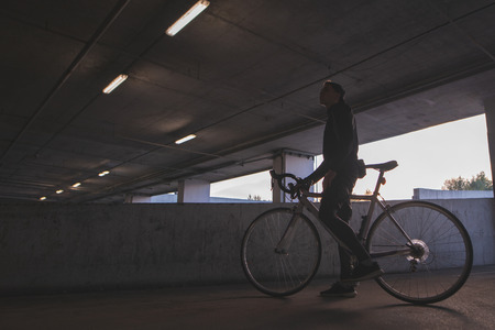An evening photo of a young cyclist standing under the bridge with a bicycle. Background with a cyclist in dark colors.の写真素材