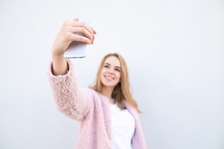 The attractive blonde in a white shirt and a pink fur coat, smiles and takes selfie on phone, white...