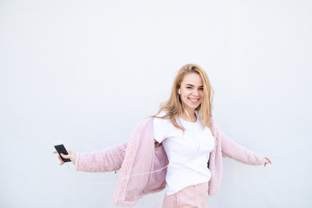 Happy, stylish girl wearing light cute clothes stumbles against the background of a white wall with a smartphone in her hands, listens to music in the headphones and smiles. Girl dances. Copyspaceの写真素材