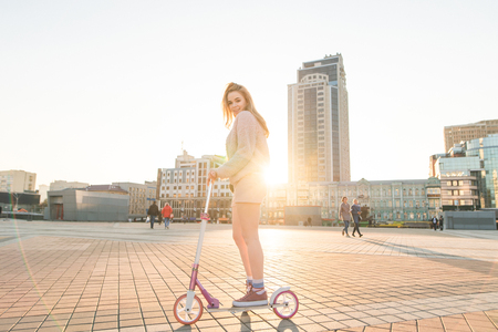 Cute girl student in pink clothes rides a kick scooter along the streets of the city at sunset. Girl rides the city on ecological transport, looks into the camera and smiles. Scooter Concept.の写真素材