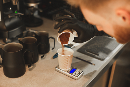 Close up photo of barista weighing coffee grains on digital scale. Hand of male barista pouring roasted coffee grains in a paper cup and weighing it before brewing coffee.の写真素材