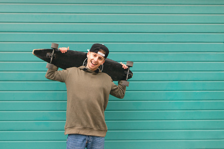 Smiling teenager in street clothes stands with a longboard in his hands against the background of a turquoise wall, looks at the camera and smiles. Copyspaceの写真素材