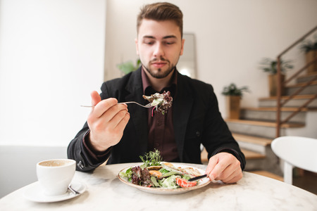Attractive man with a beard eating a diet salad in a cozy restaurant. Man took the green salad on the fork. Focus on the green on the fork. Good food.の写真素材