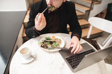 Business man sitting with a laptop in a cafe and eating salad, a cup of coffee on the table. Busy man dishes in a cafe with healthy food and works on a laptop.の写真素材