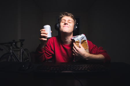 Satisfied young man in the headset smiles and holds a drink with snacks in his hands. Gamer sits at the computer, dishes snack and gets pleasure.の写真素材