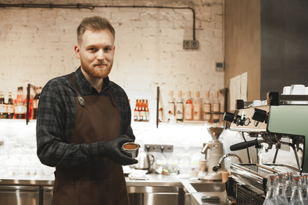 Smiling bearded coffee shop owner is ready to prepare coffee with a coffee machine. Cheerful cafe owner is holding a portafilter and making a cappuccino and espresso at his cafe.の写真素材