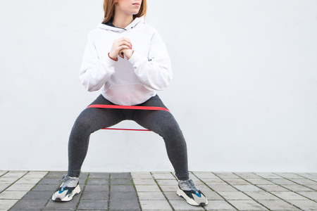 Close-up fitness girl squats with a rubber band on a white background. Enjoy sports on the street. Warm up before running. Copyspaceの写真素材