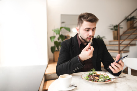 Attractive man in a suit sits in a cozy restaurant, eating salad with a plate and using a smartphone. Business man use a smartphone during lunch in the restaurant. Focus on the smartphone. Copyspaceの写真素材