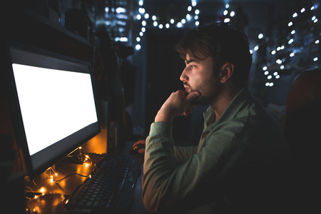 Man with a beard sits at home in a room, but uses a computer. Man works at night on a computer, looks at the screen with a serious look.の写真素材
