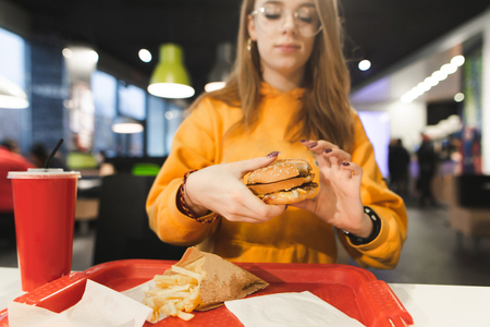 Girl teenager with a fast food tray sitting at a restaurant table, holding a burger. Focus on a burger in the hands of a girl who eats fast food. Student and fast foodの写真素材