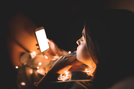 Woman lays on a bed at night, holds a smartphone in her hands, typing a message on her phone, uses the internet.Girl in the luminous flames holds the smartphone in her hands before bed.の写真素材