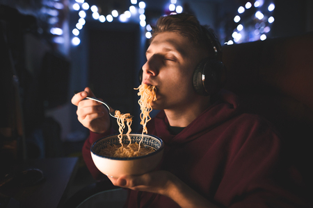 Portrait of a teenager in headphones sitting at a table near a computer and eating a night-time noodle soup in a room. Young man dinner with a noodle soup, uses a computer.の写真素材