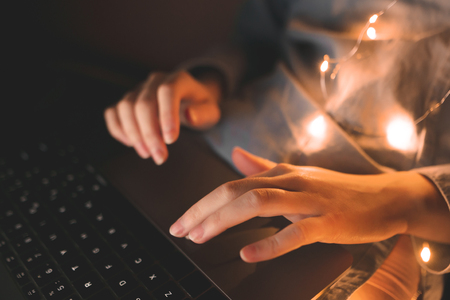 Close-up photo of feminine hands is working on a laptop at night. Woman with a laptop on her legs is lying in a bed of lights and uses a laptop. Focus on the hand touches the touchpad.の写真素材
