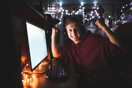 Portrait of an emotional teenager sitting at home on a computer and rejoices in victory with his hands raised. Joyful teenager plays video games at home on the computer, looks at the camera and smilesの写真素材
