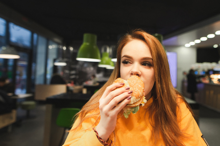 Sweet girl holds a large tasty burger with two hands, bites and looking sideways in close-up. Close-up portrait of a girl eats a burger, fast food at the restaurant background. Copyspaceの写真素材