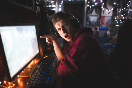Amazed young man enjoys a camping at night at home, shows his finger on the screen and looks at the camera with astonished look. Gamer plays video games on a computer.の写真素材