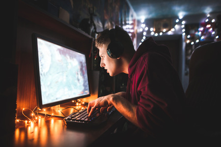 Concentrated teenager sits at a computer table in the cozy room, looks at the monitor and plays video games. Student gamer plays games at night on a computer. Night gamingの写真素材