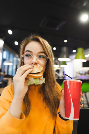 Portrait of an amusing pretty girl who eats a tasty burger and holds a glass of beverage in her hands. Attractive girl eats fast food in a light fast food restaurant. Girl eats a burgerの写真素材
