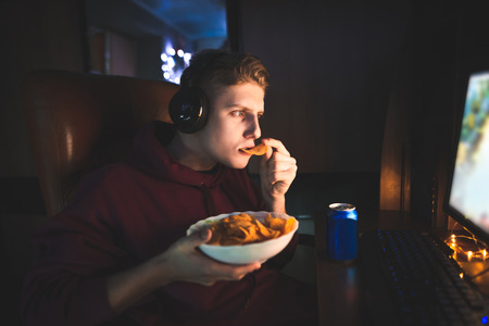 Young man eats chips at night, drinks beer and uses a computer. Portrait of a gamer playing video games on a computer and eating snacks. Supper at the computer at night.の写真素材