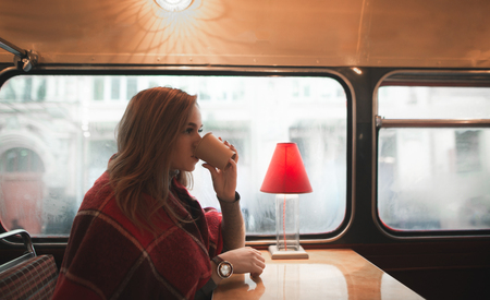 Winter portrait of a beautiful woman who drinks coffee in a cozy coffee shop. Girl is covered with a blanket sitting in a cafe with a cup of coffee in her hands and looking to the sideの写真素材