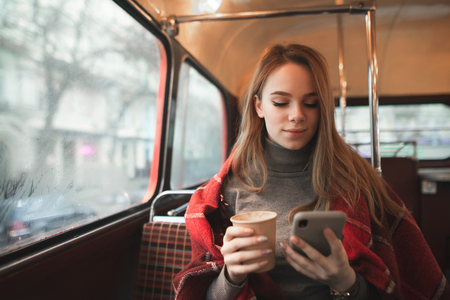 Portrait of an attractive girl covered with a blanket sits a cozy cafe with a cup of coffee in her hands and looks at the smartphone screen. Girl drinks coffee and uses a smartphone in a coffee shop.の写真素材