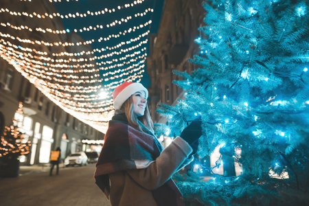 Christmas portrait of a smiling girl in warm clothes , evening on a decorated street in the lights near the Christmas tree.Girl in a holiday hat walks along the street decorated before Christmasの写真素材