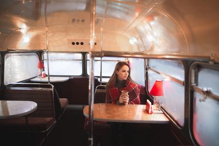 Beautiful woman wrapped in a blanket, sitting in a coffee shop with an original retro interior. Girl is sitting at the cafe bus,holds a cup of coffee his hands and warmes, looking in the windowの写真素材