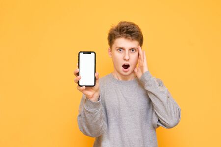 Portrait of a surprised young man in casual clothing holds in his hand a modern smartphone with a white screen and looks shocked at the camera, isolated on a yellow background. Copyspaceの写真素材