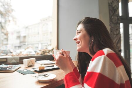 Happy girl sitting at the table in a cafe with a dessert and a cup of delicious coffee in her hands,...