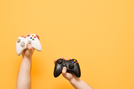 Gaming background. Two gamepads in the hands of a man on a yellow background. White and black joystick holds, isolated. Copyspaceの写真素材