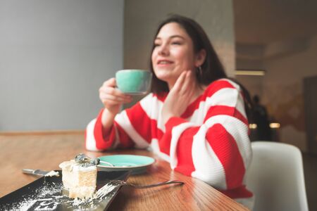 Focus on a plate with a delicious cheesecake, on the background of a girl. Smiling girl sits in a cafe with a cup of coffee in her hands and a plate of appetizing cake on the table.の写真素材