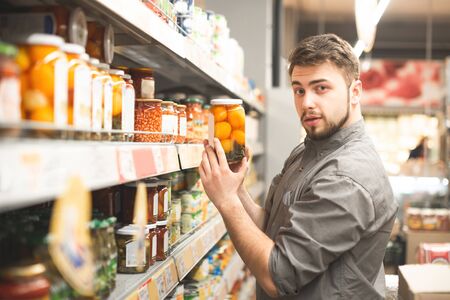 Portrait of a beautiful man with a beard standing in the aisle of the supermarket,a department of canned food,holding a jar of canned vegetables and looking into the camera.Man buys canned vegetablesの写真素材