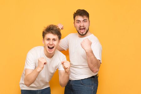 Portrait of two men winners, rejoicing with their hands raised, looking into the camera, wearing a white T-shirt, on a yellow background. Two happy friends rejoice.の写真素材
