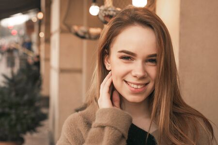 Close portrait of an attractive stylish girl wearing a beige coat, looking into the camera and smiling against the backdrop of the building. Street close up portrait of a happy girl in spring clothes.の写真素材