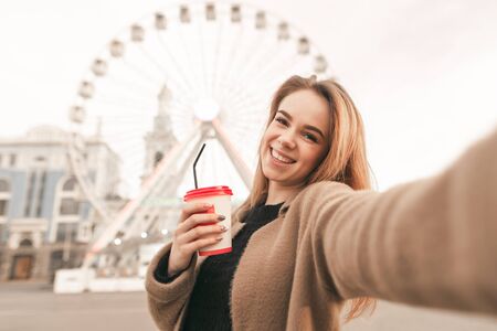 Attractive blonde girl in casual clothing, wearing a coat, holding a cup of coffee in his hands, takes selfie, looking into the camera. Stylish girl takes selfie, background of the ferris wheelの写真素材