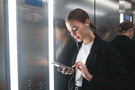 Smiling businesswoman is using her smartphone with headphones in the elevator. Cheerful office worker is texting on the phone and listening to music in her earphones.の写真素材