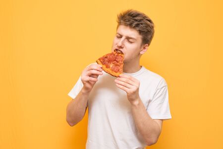 Guy in a white T-shirt eats an appetizing piece of pizza with his eyes closed on a yellow background and gets pleasure. Young man bites a pizza, isolated on a yellow background.の写真素材