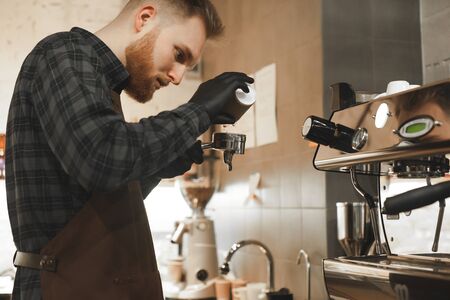 Coffee shop owner is pouring coffee into the portafiler at the coffee machine. Bearded barista is holding a holder at the cafe and brewing coffee.の写真素材