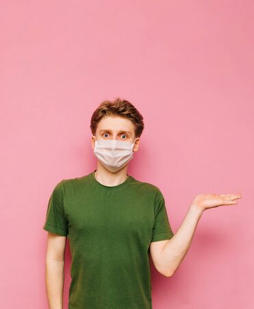 Portrait of a surprised young man in a gauze mask on a pink background, looking at the camera and holding a place for advertising. Coronavirus pandemic. COVID-19. Quarantine.の写真素材