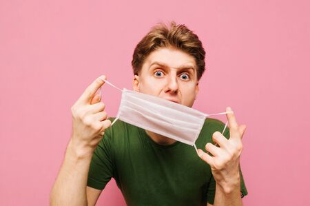 Close-up portrait of an angry guy wearing a medical mask on his face on a pink background, looking at the camera with a crazy face. Trained emotional guy removes a gauze mask from his face.の写真素材