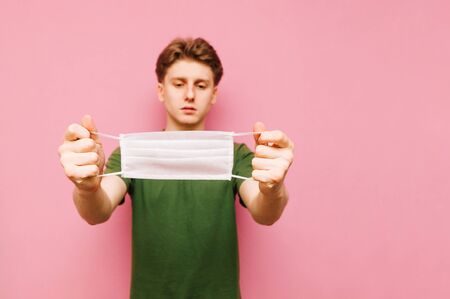 Handsome man in bright clothes stands on a pink background with a medical mask in his hands. Focus on gauze mask in the hands of guy, puts on for protection. Coronavirus pandemic. Quarantine.の写真素材