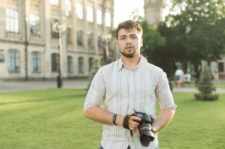 Professional bearded photographer stands in the park with a camera in hand, looking in camera with a serious face.Street photo of handsome man with camera in hands on background of university buildingの写真素材