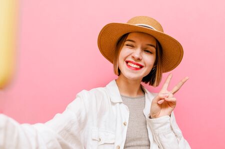 Happy tourist girl in sun hat and summer clothes makes selfie on smartphone on pink background,...