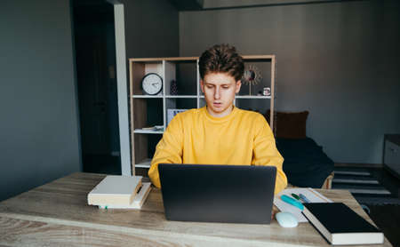 Young man sits at home with a laptop and books and studies. Student doing homework at home at the table in the bedroom. Distance Learningの写真素材
