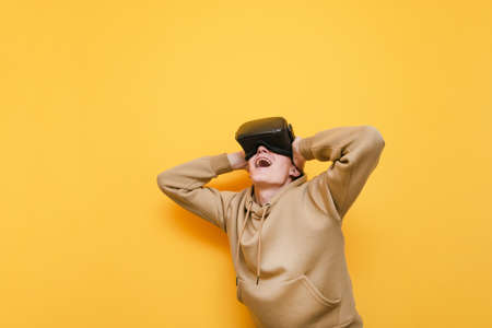 Joyful young man in VR helmet stands on yellow background, looks up with surprised face and mouth...