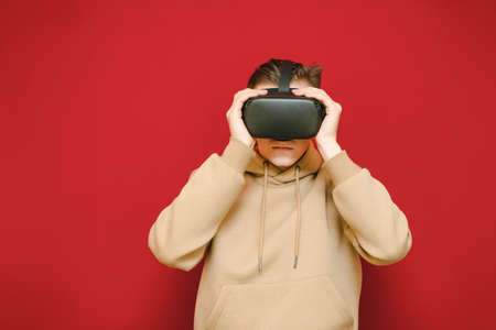 Young man in a beige sweater is wearing a virtual reality helmet on a red background. Portrait of a guy in VR glasses. Isolated. VR gaming concept.の写真素材