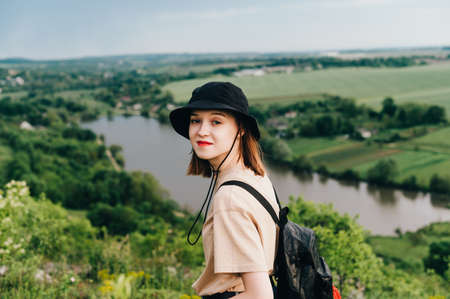 Positive girl tourist in casual clothes standing on a hill against the backdrop of the lake and...