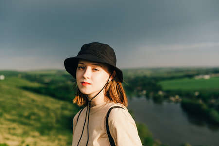 Close portrait of an attractive girl tourist in casual clothes standing on a mountain against the backdrop of a lake at sunset, and looking at the camera with a serious face. Domestic tourismの写真素材