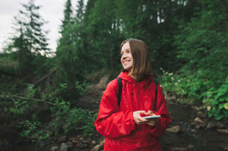 Young girl hiker in a red raincoat is smiling holding phone in her hands on a background of mountain...