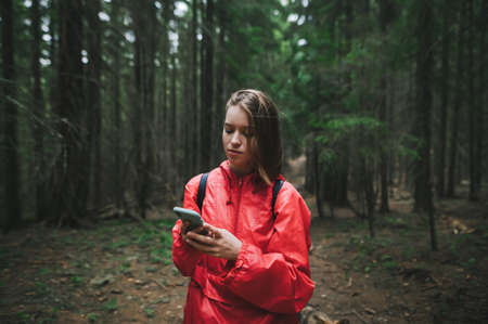 Beautiful young hiker girl in a red raincoat is texting using her phone in the mountain forest....
